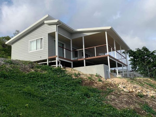 Gray house with a porch on a hillside with green grass and a cloudy sky.