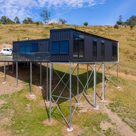 Black container home raised on stilts, hillside setting, with clear glass balcony.
