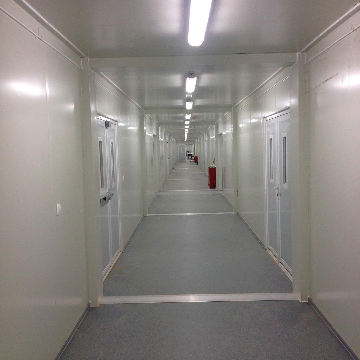 Long, sterile hallway with white walls and doors, bright fluorescent lighting, and a grey floor.
