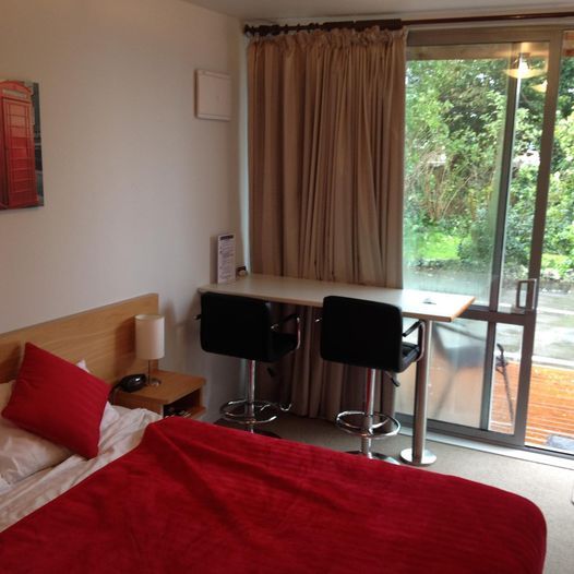 Bedroom with a bed, small table, two bar stools, and a sliding glass door with a view of greenery. Red accents.