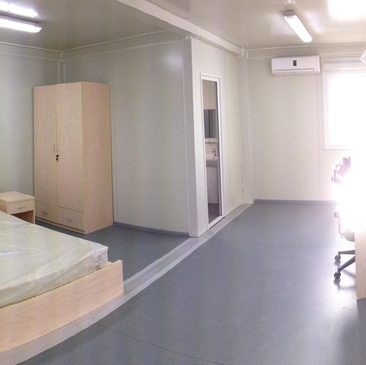Empty room with bed, closet, and small bathroom visible. Grey floor and off-white walls.