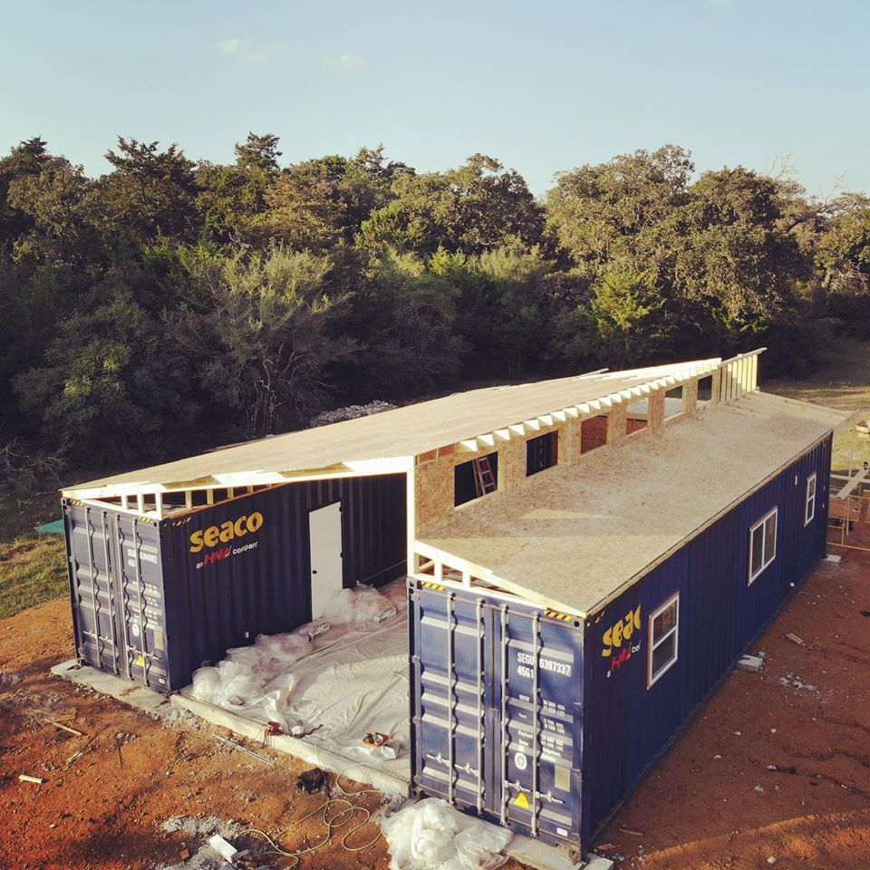 Two dark blue shipping containers connected under construction with a wooden roof, outdoors.