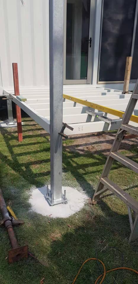 Construction of a deck. A metal post is set in concrete, attached to beams and near a building.
