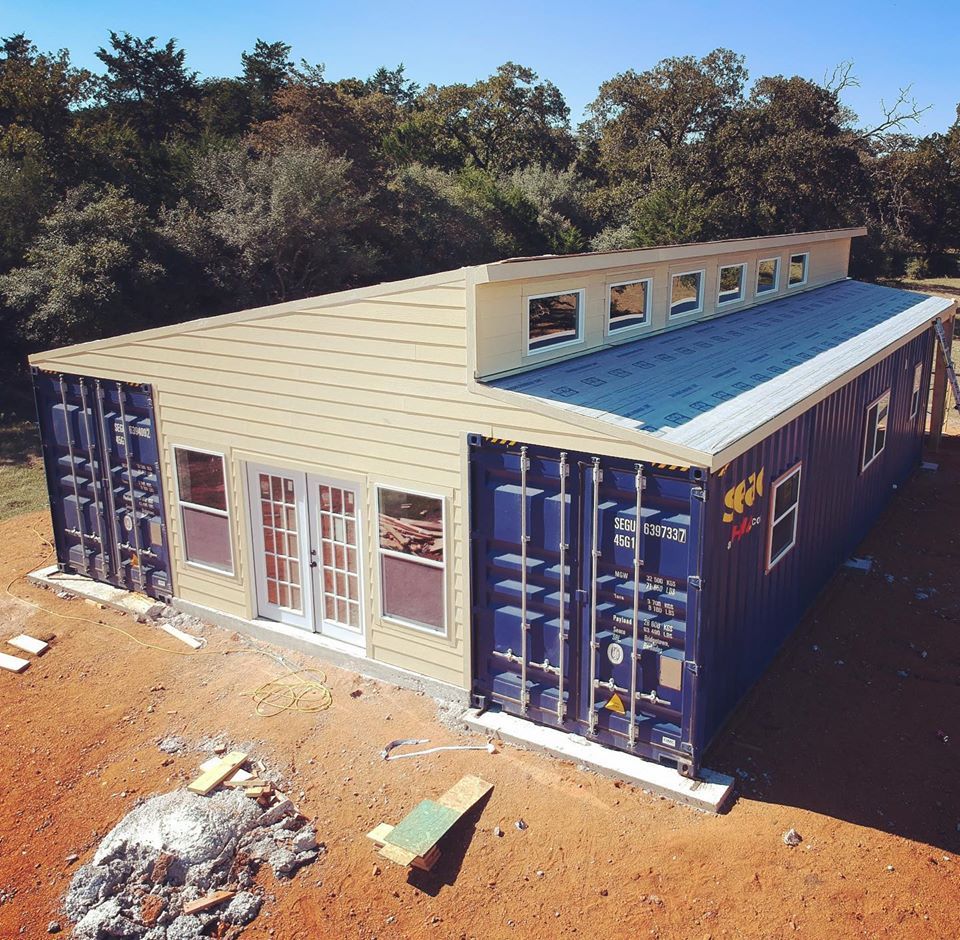 Shipping container home with blue walls, beige roof, windows, and double doors. Set in a wooded area.
