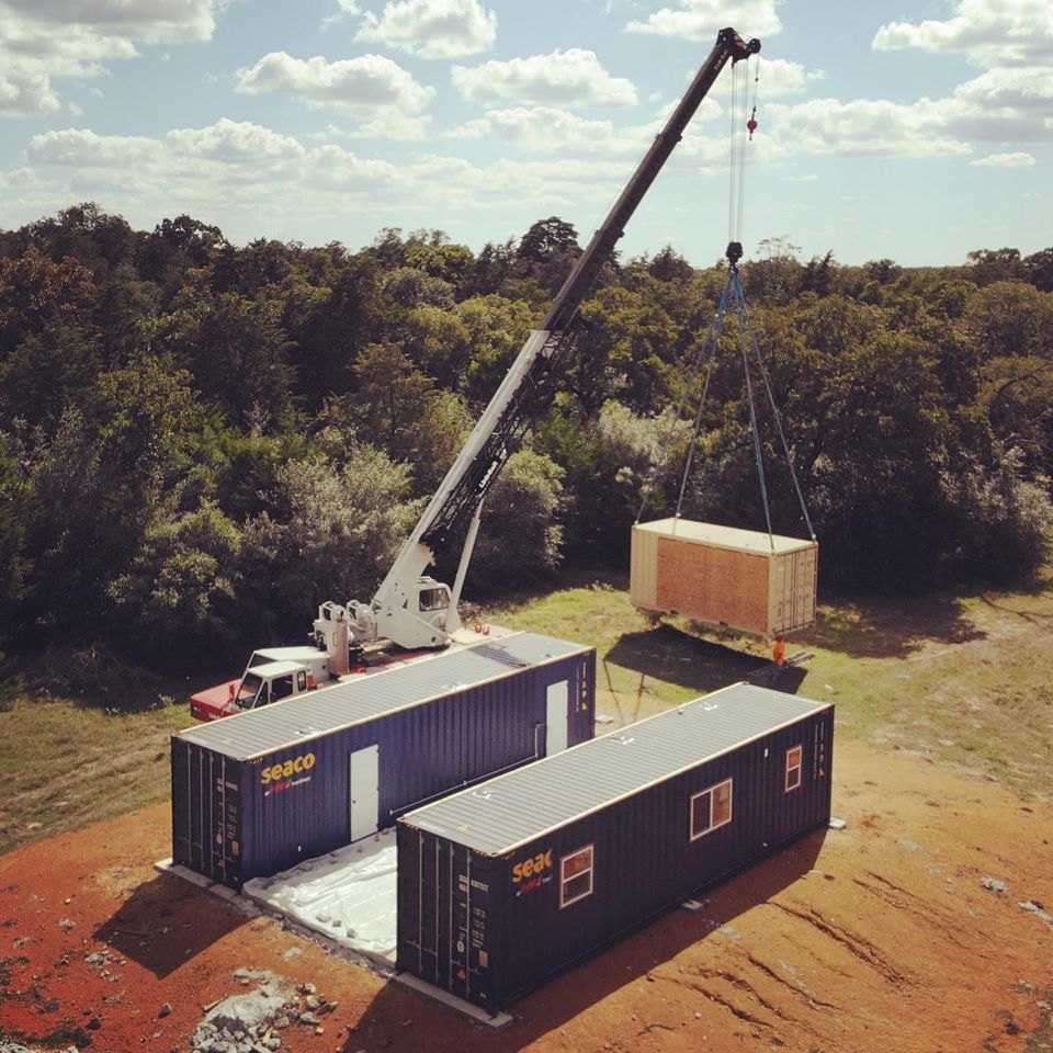 A crane lifts a wooden structure above two shipping containers on a red dirt field, surrounded by trees.
