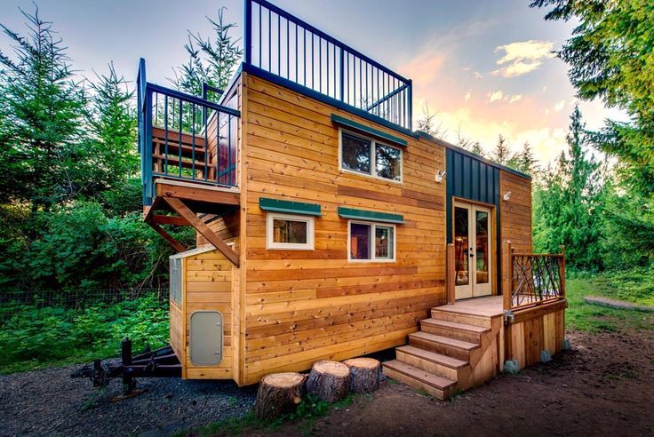 Tiny wooden house on wheels, with steps, deck, and balcony, set in a forest.