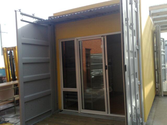 Shipping container converted into a structure with yellow exterior, sliding glass doors, and open gray doors.