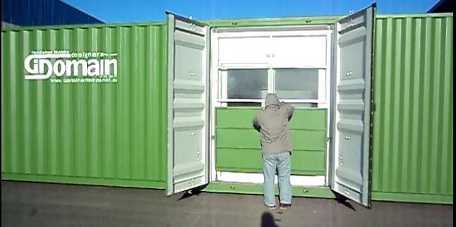 A person stands in front of a green shipping container. The container's doors are open to reveal a window.