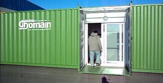 A person enters a green shipping container that has been converted into a building, with the door open.