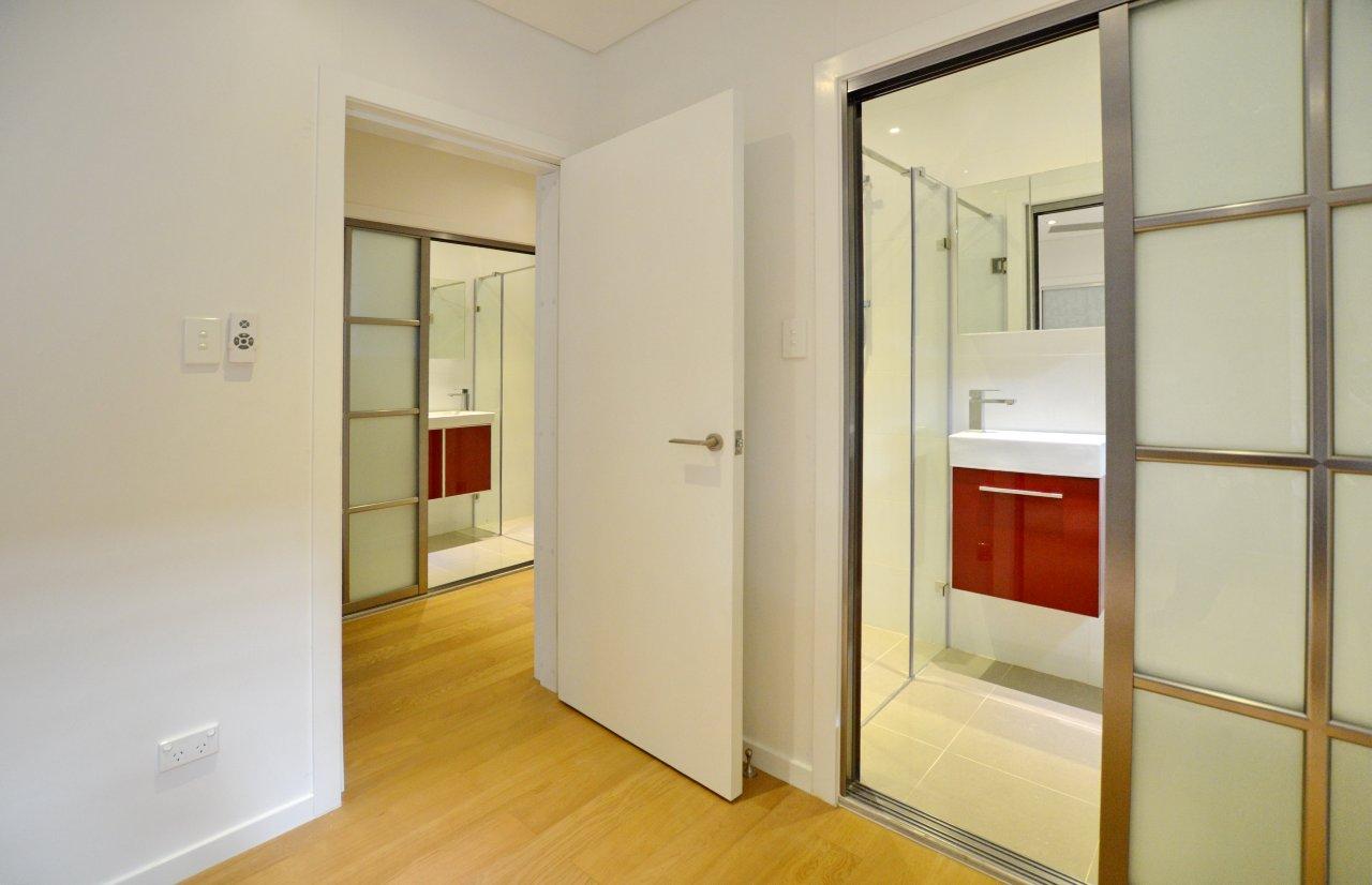 Interior view of a room with two bathrooms visible. Bathrooms have red vanities, glass doors, and light wood floors.