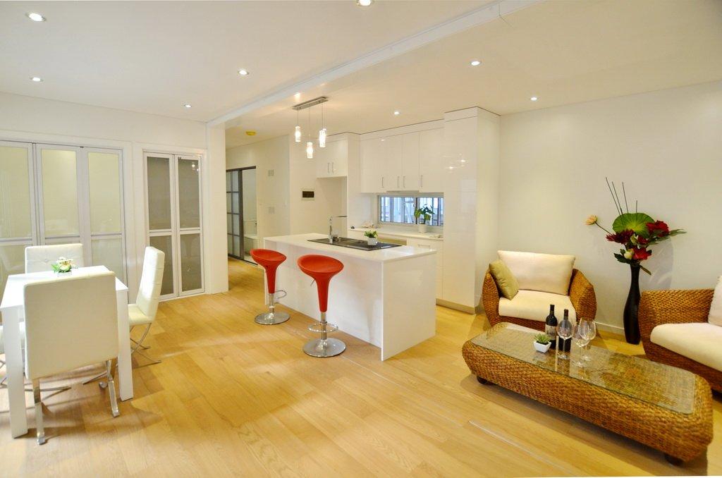 Open-plan living space: white kitchen island with red stools, dining area, wicker furniture, hardwood floor.