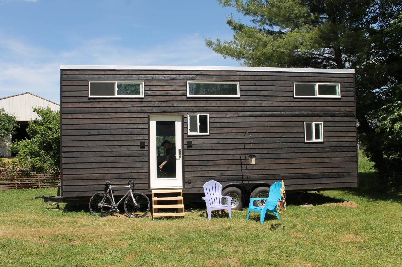 Tiny house on wheels with black siding, door, windows, bike, and chairs in a grassy yard.