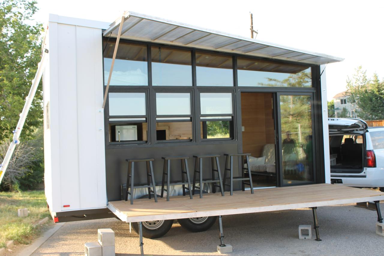 Tiny house on a trailer with an extended deck, large windows, and an awning.