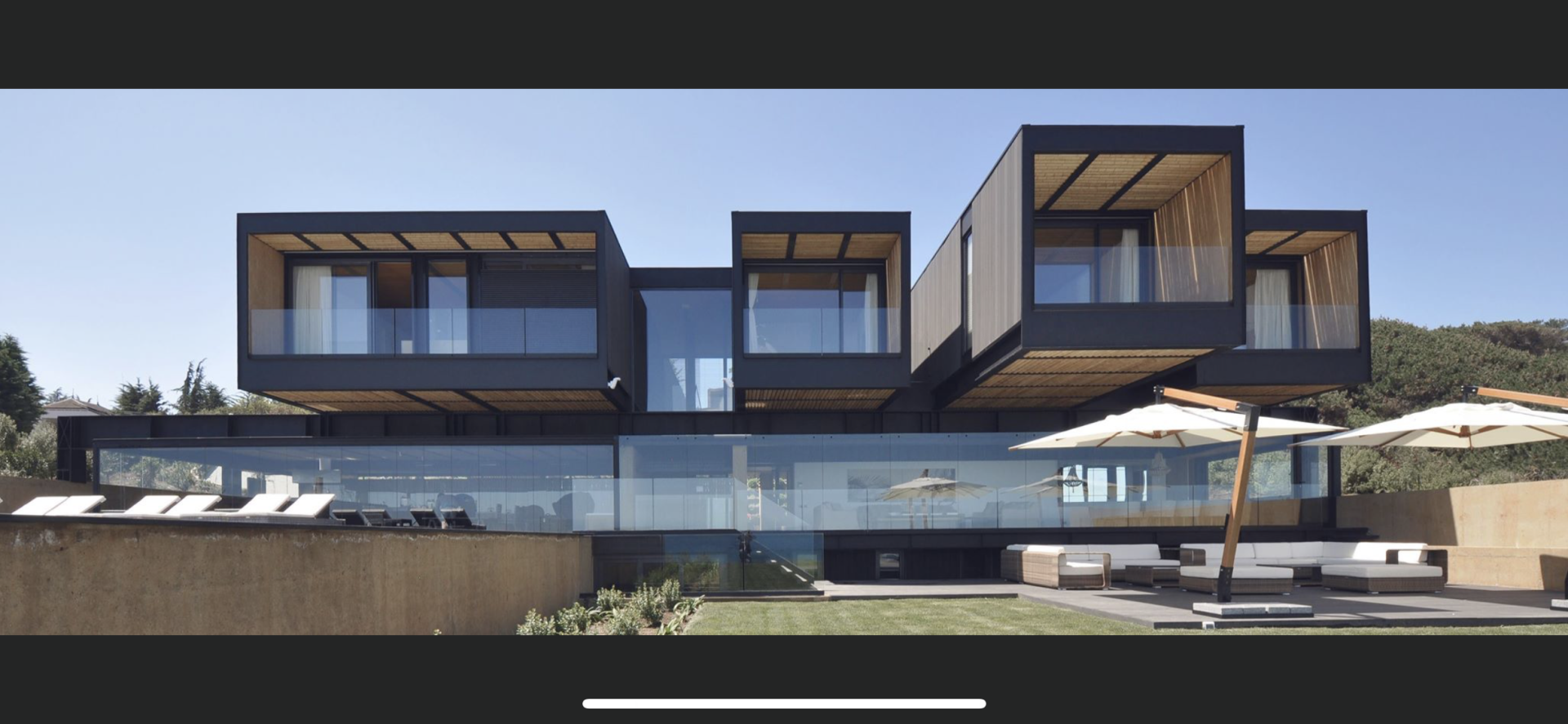 Modern home with black rectangular structures over a glass-walled base, outdoor seating, and umbrellas.