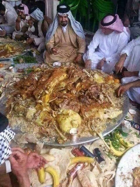 People in traditional white robes eat a large platter of food, possibly in a communal setting.