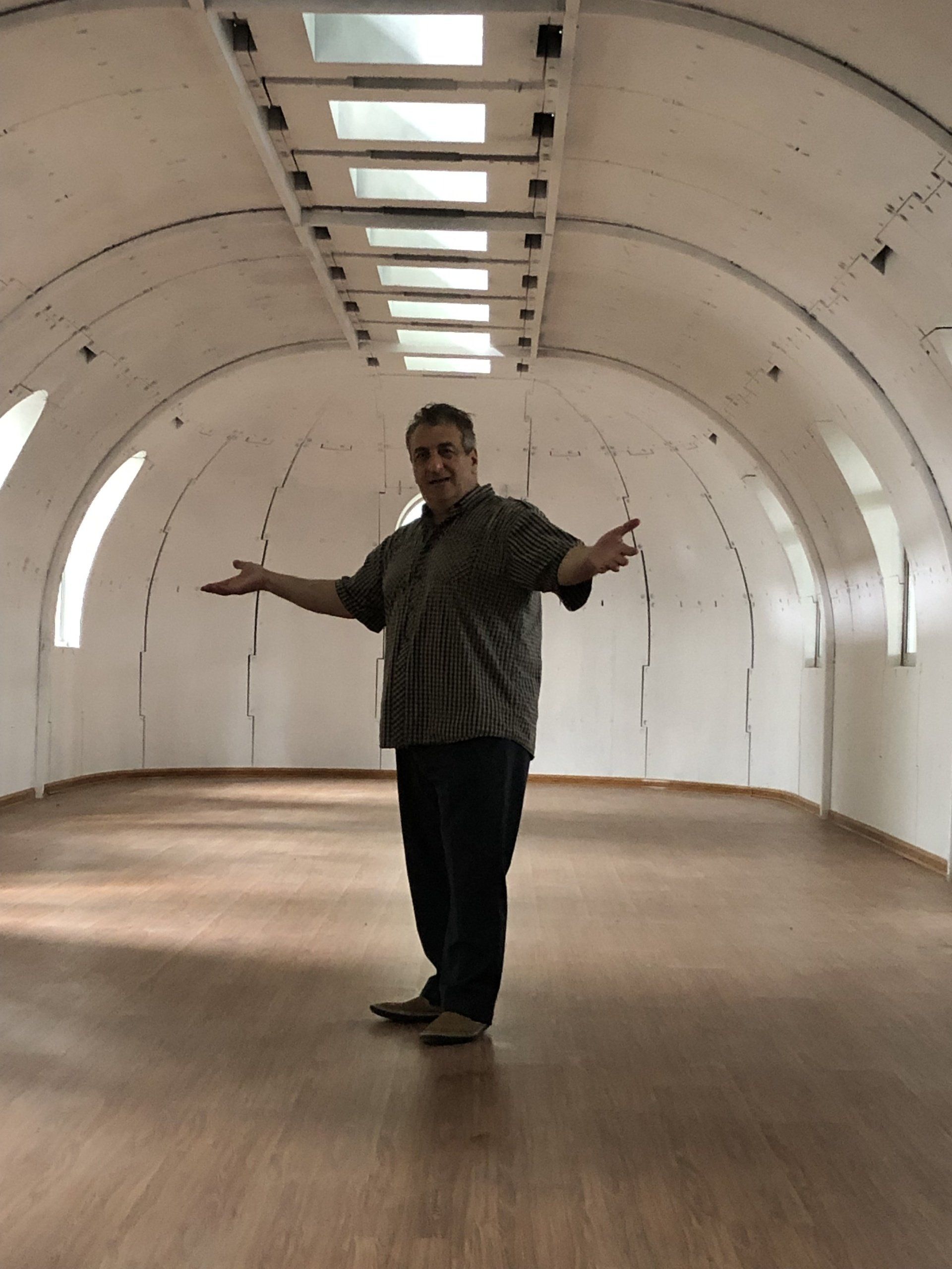 Man stands with arms open in a white, arched-roofed room with wooden floor.