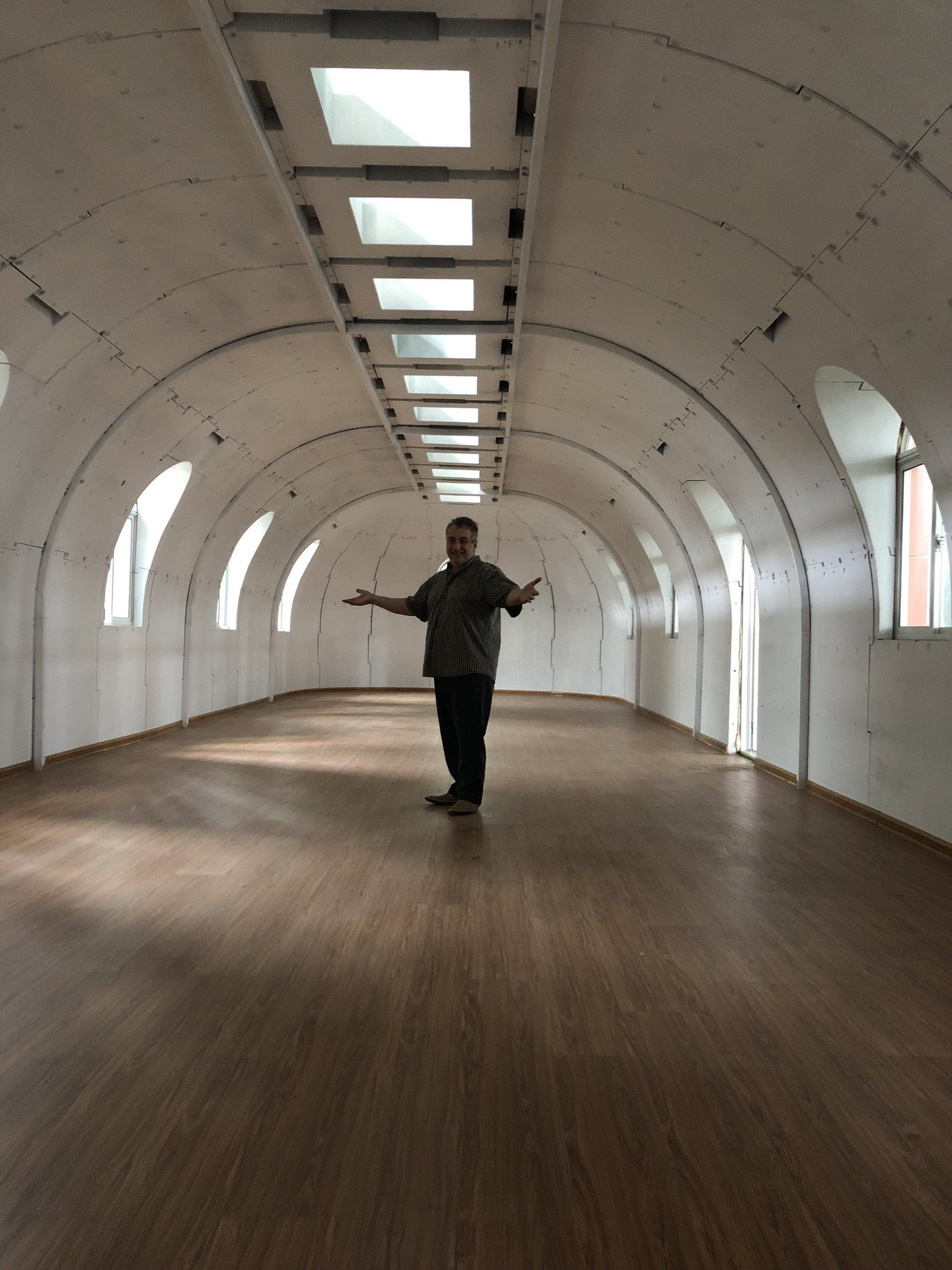 Man standing in a long, arched room with a wooden floor, arms outstretched, natural light from windows and skylights.