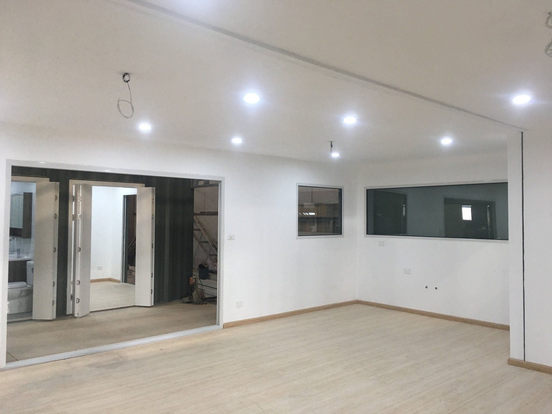 Empty room with white walls, recessed lighting, and wooden floors. Two rectangular windows and open doors.