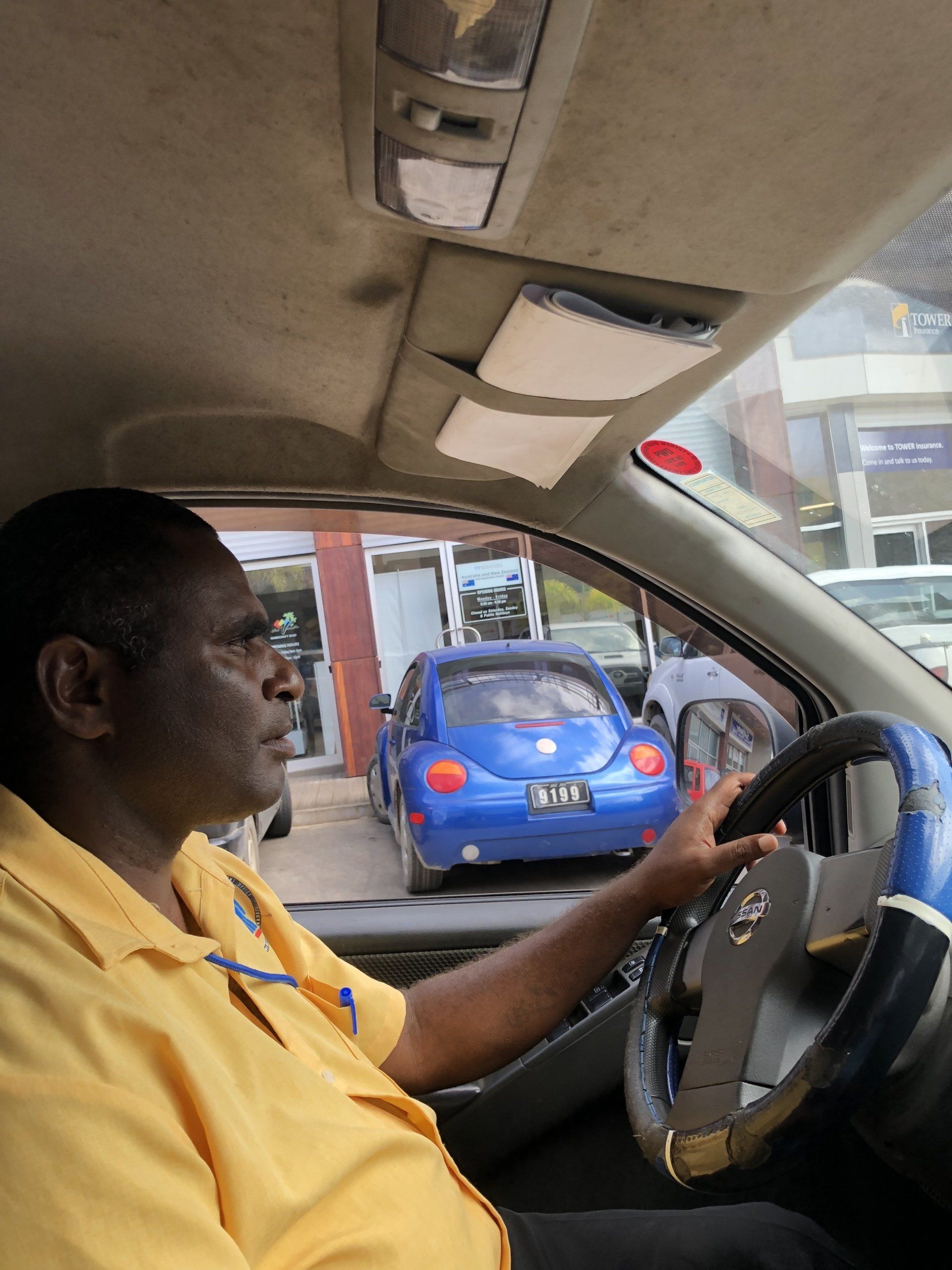 Man driving a car, hands on the steering wheel, looking forward. Blue car visible in front.