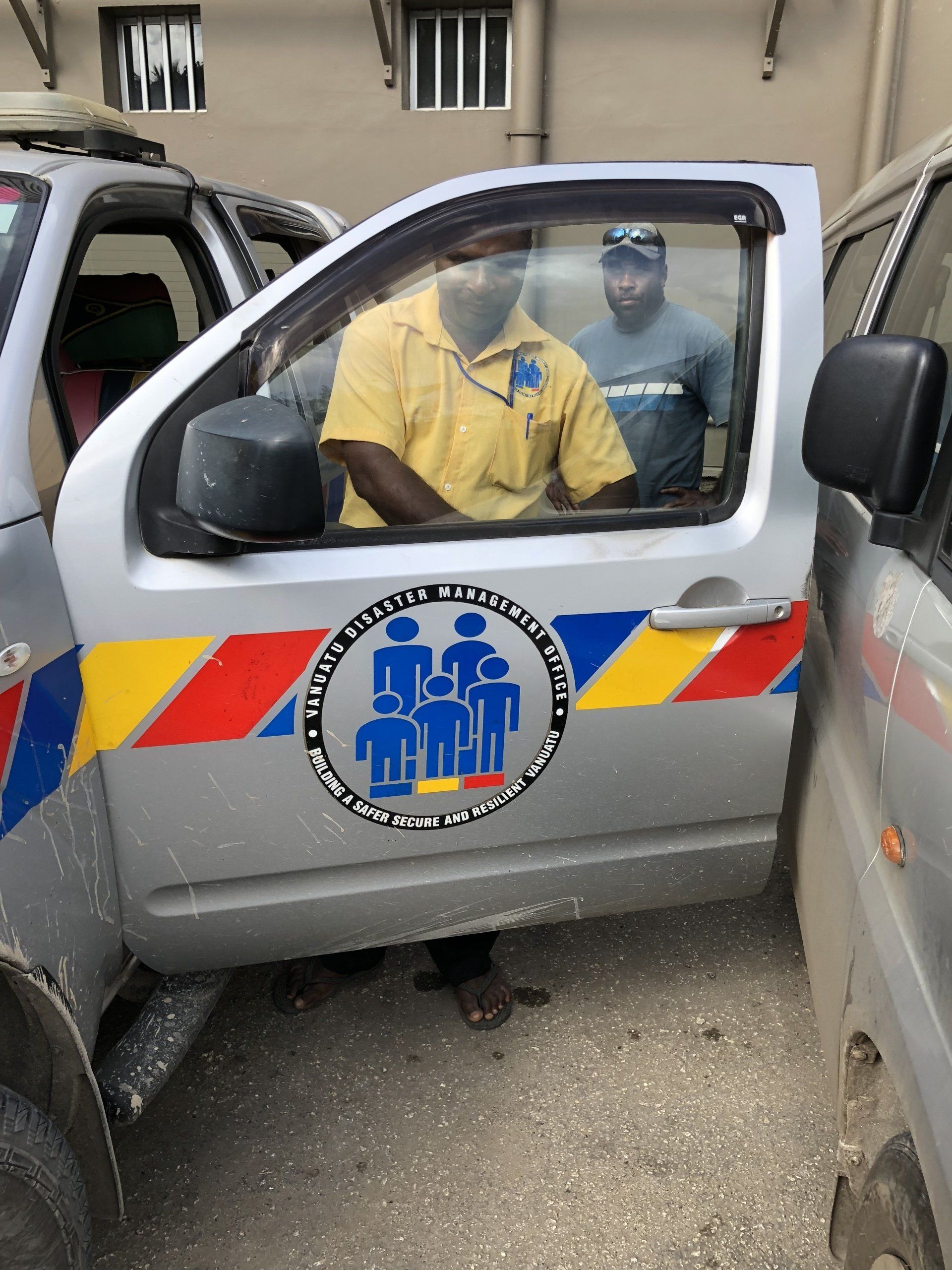 Man in yellow shirt in a silver vehicle with the logo of an organization. Another man in the background. Door open.