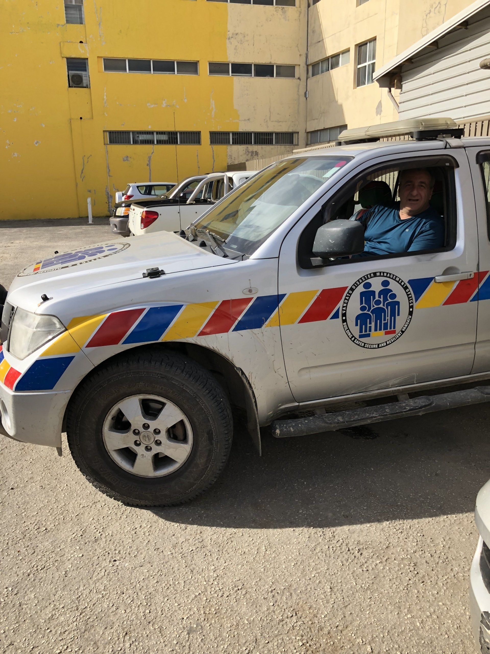 Man in a silver and colored-stripe service vehicle parked outside a yellow building.