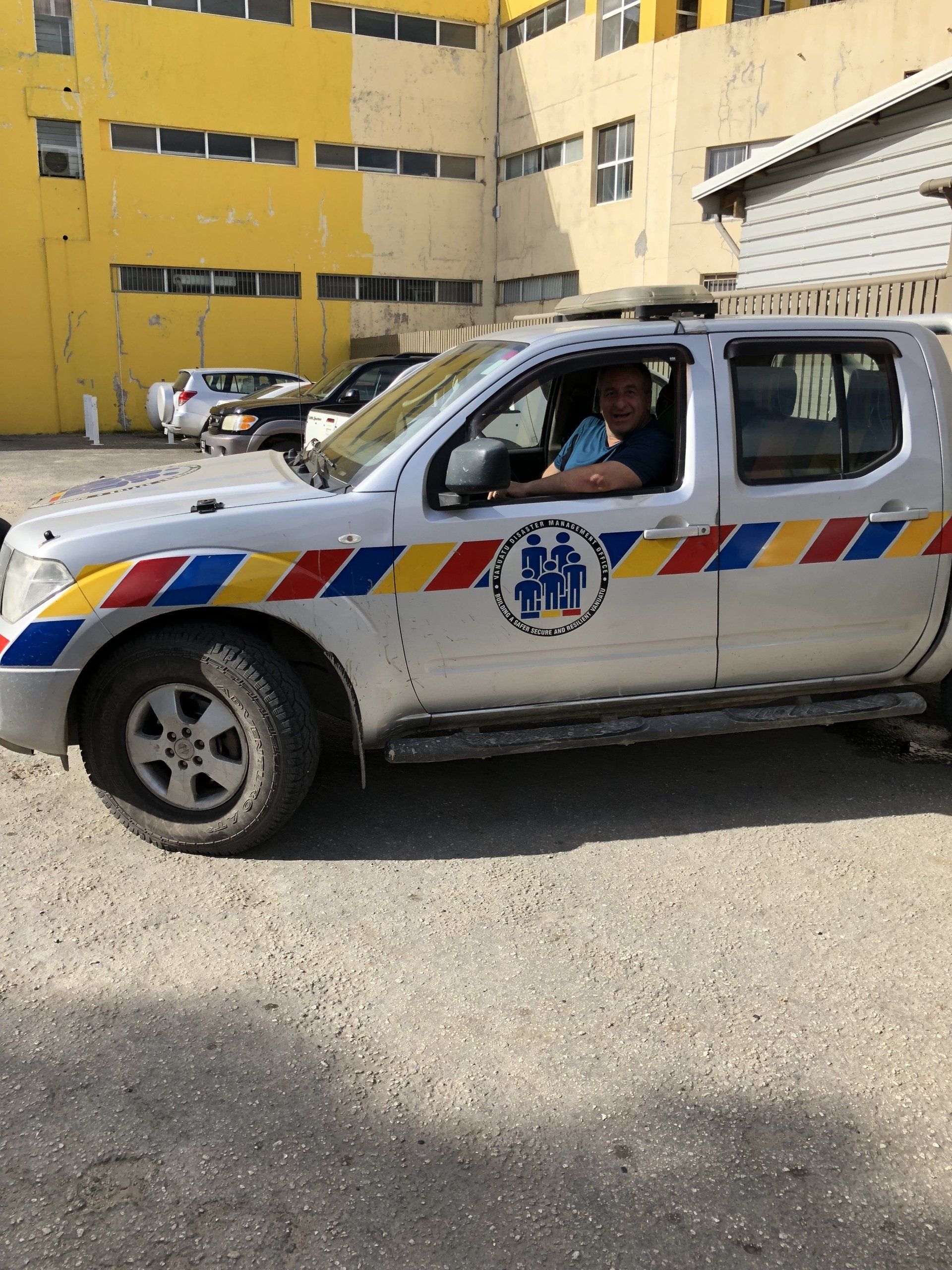Silver pickup truck with logo and color stripes; man in driver's seat; yellow building in background.