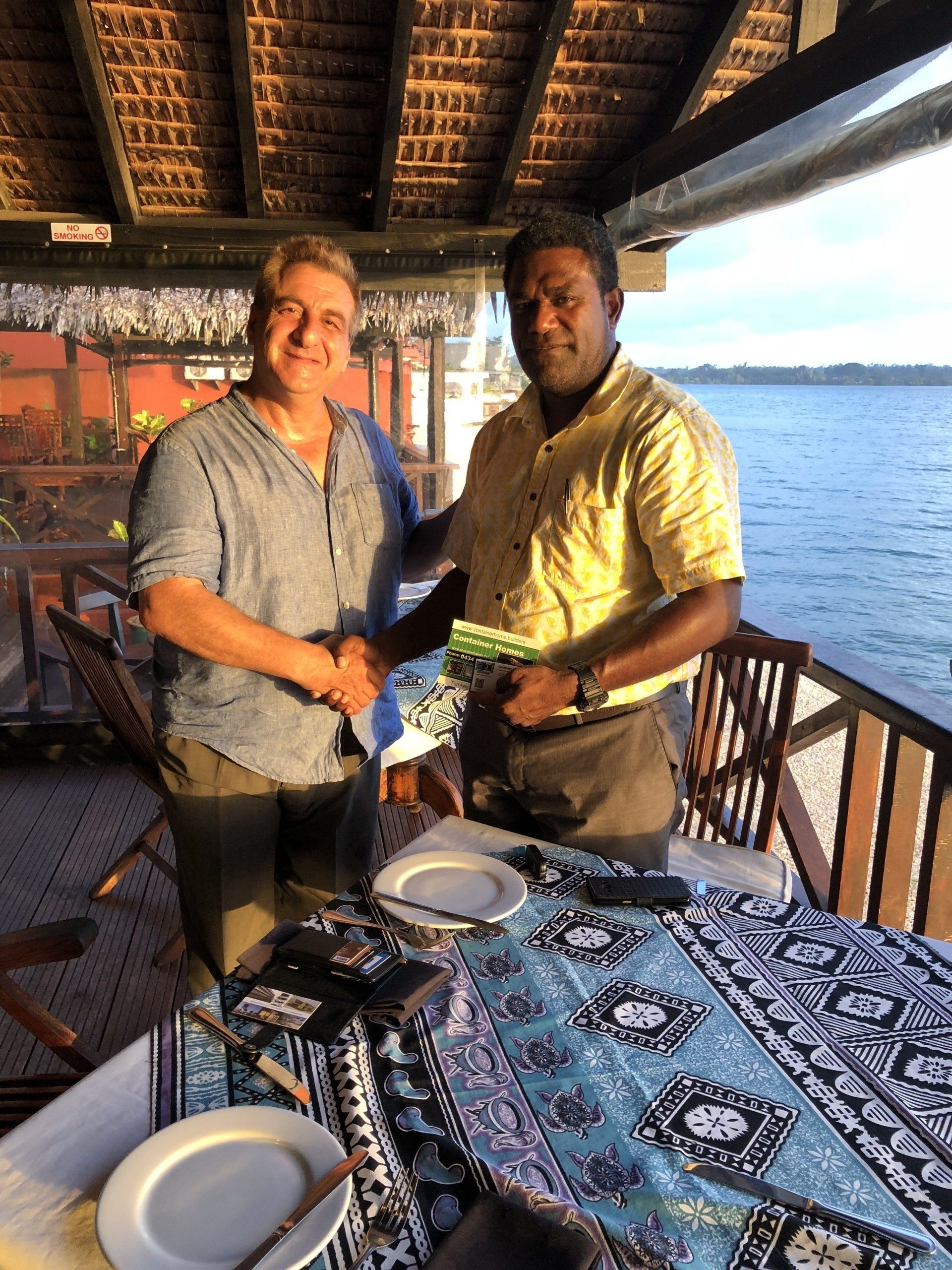 Two people shaking hands near a table set with dishes and a patterned cloth, overlooking a body of water.