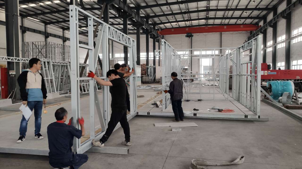 Workers assembling a metal frame structure in a factory. White walls, various tools, and overhead cranes are visible.
