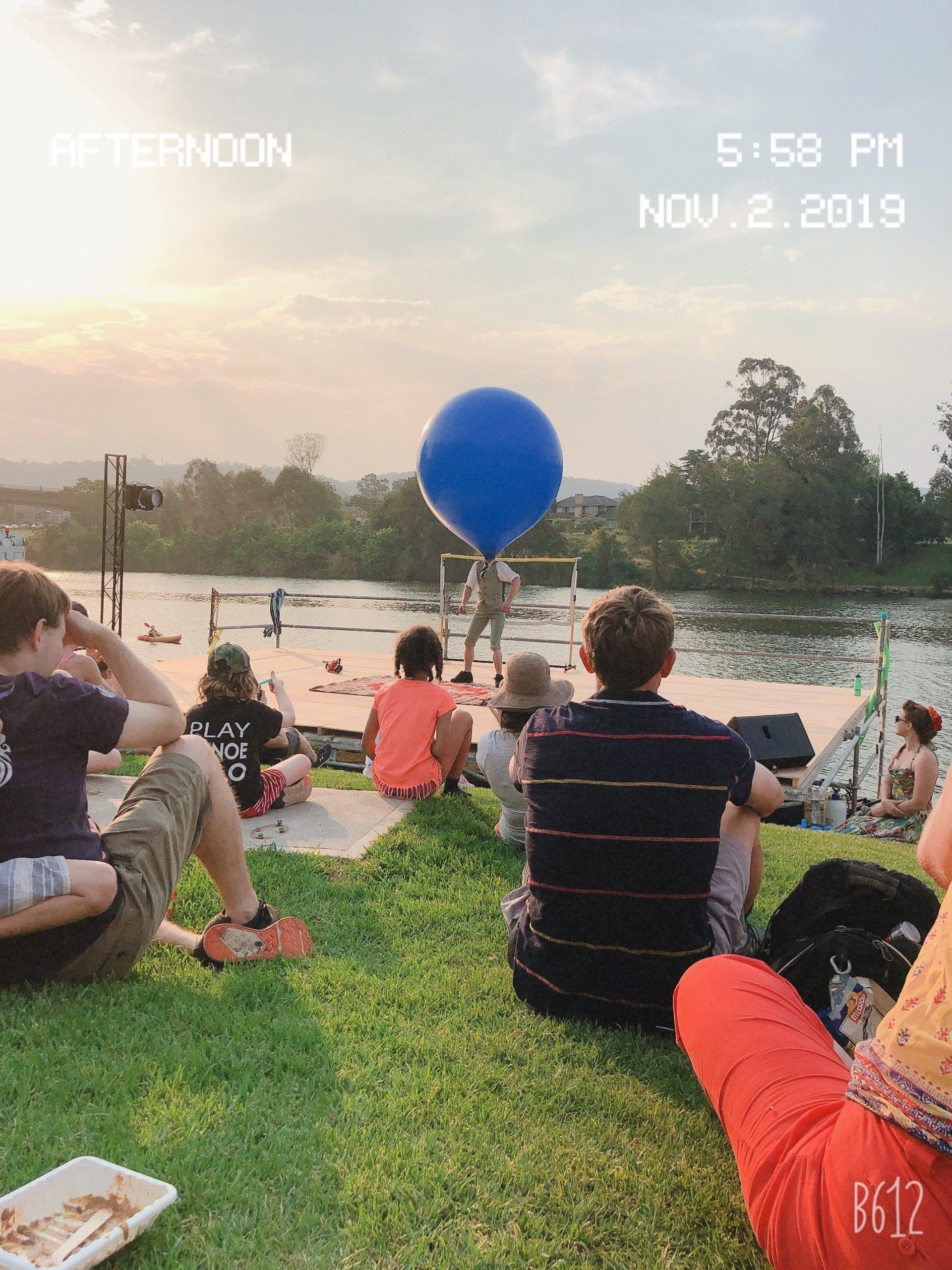 People watching a blue balloon performance near a body of water at sunset.