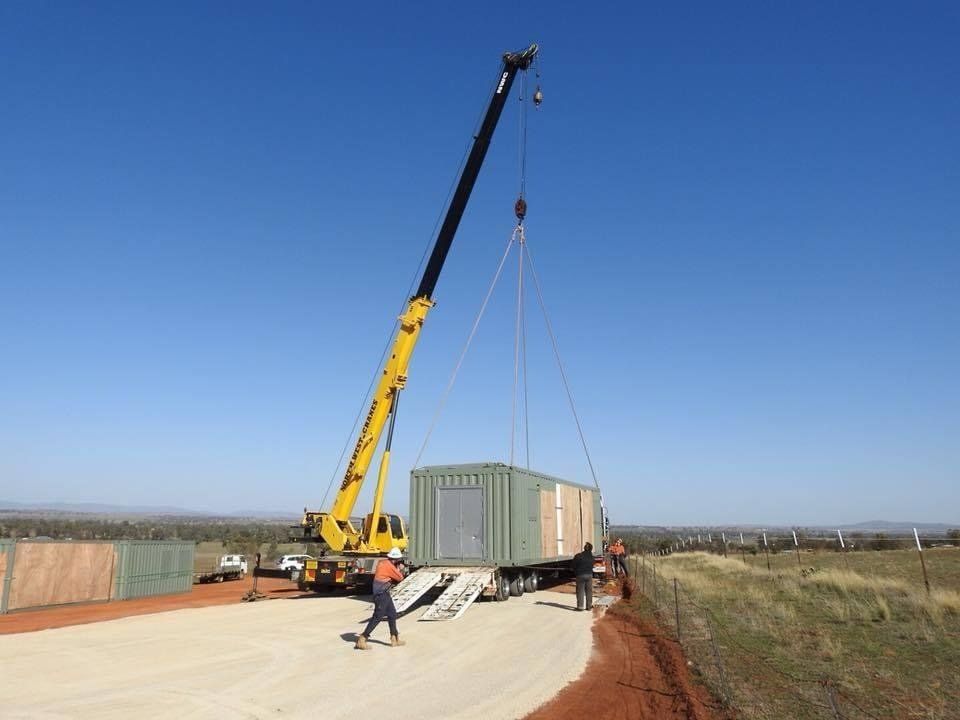 Crane lifting a container from a truck on a dirt road. Workers and a vehicle are visible. Blue sky.