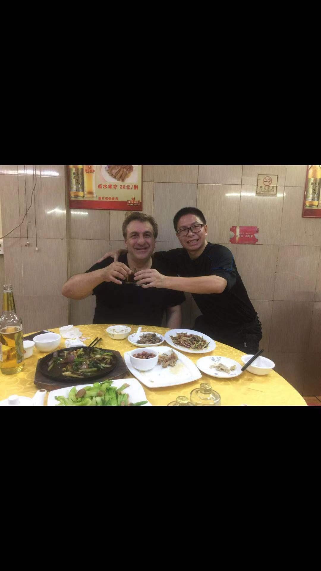Two men toasting drinks at a table in a restaurant. Food and drinks are on the table.