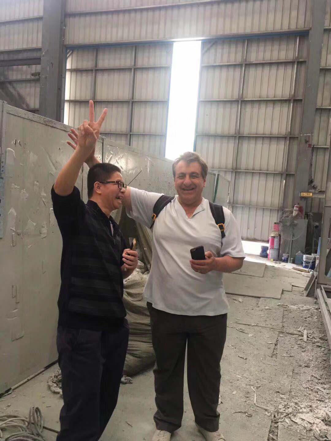 Two men in a factory; one raises a peace sign, the other holds a phone and smiles.