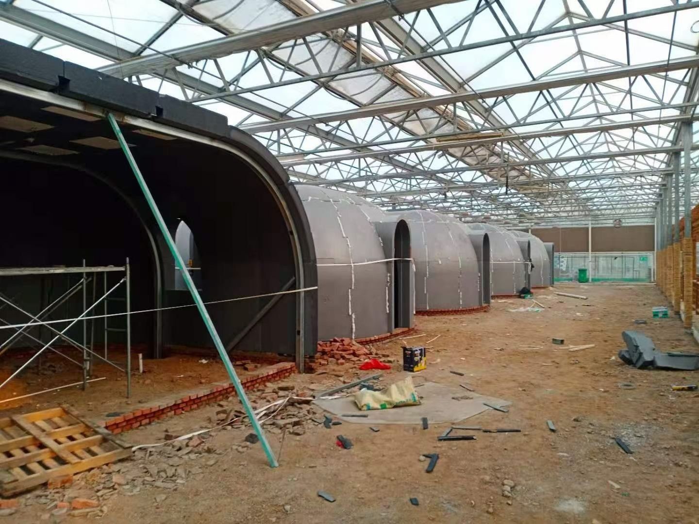 Interior of a greenhouse under construction with several curved structures. Scaffolding, debris, and exposed framing are visible.