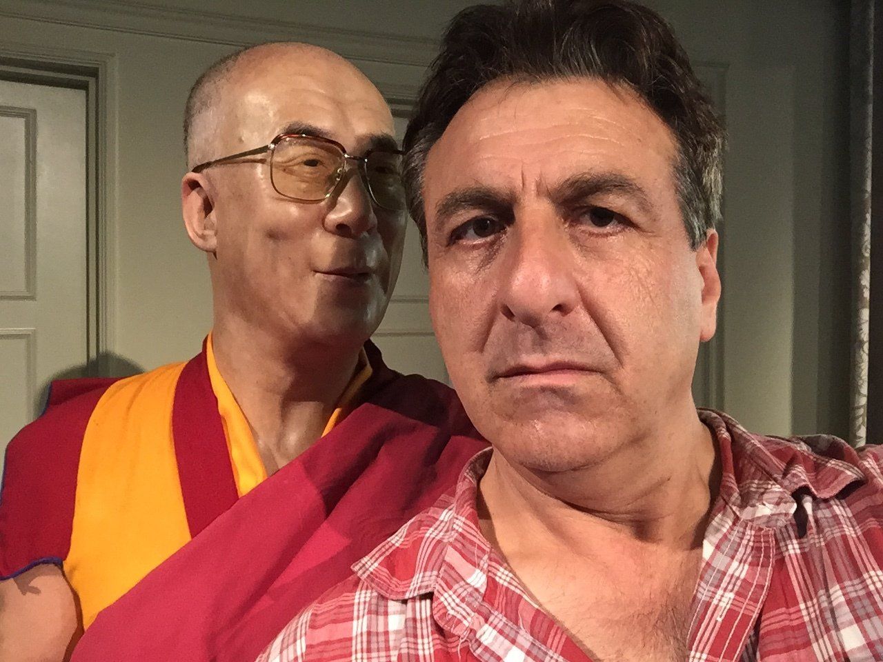 Man taking a selfie with a wax figure of the Dalai Lama. Man in plaid shirt, Dalai Lama in traditional robes.