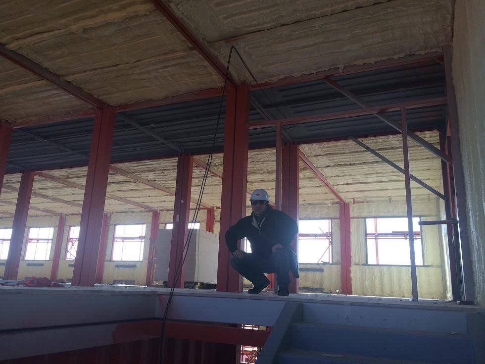 Construction worker squats, observing a building interior with exposed beams, spray foam insulation, and windows.
