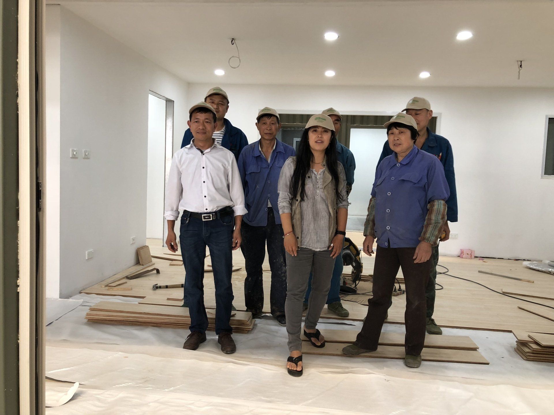 Group of people in construction site; workers in blue shirts and hats, woman in silver top.