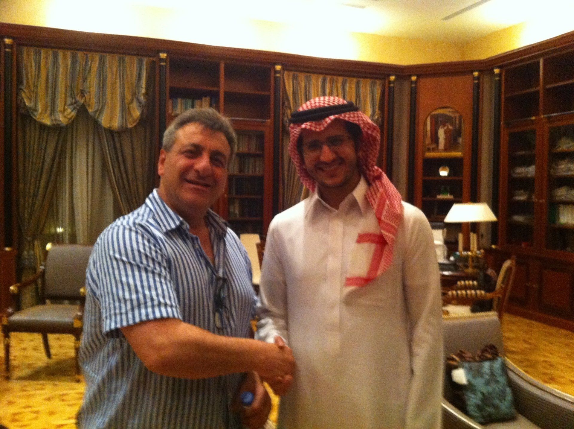 Two men shaking hands in a wood-paneled room. One in Western clothes, the other in traditional Arab attire.