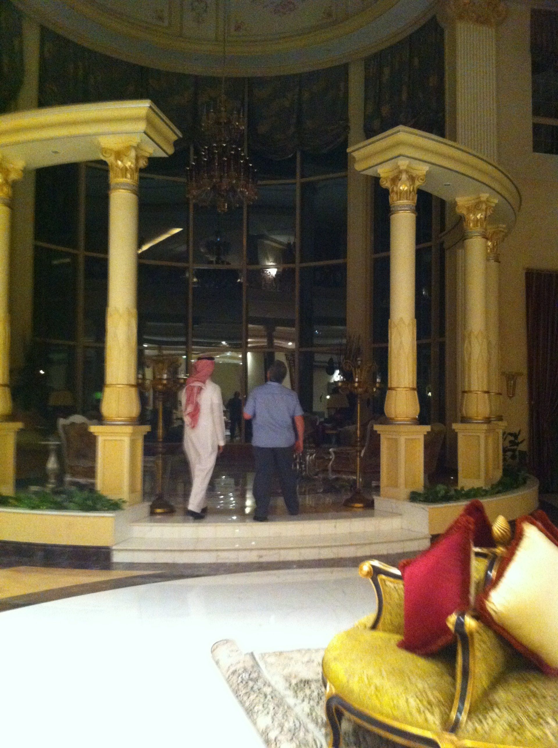 Two men walking inside a large building with ornate columns, glass windows, and luxurious furniture.