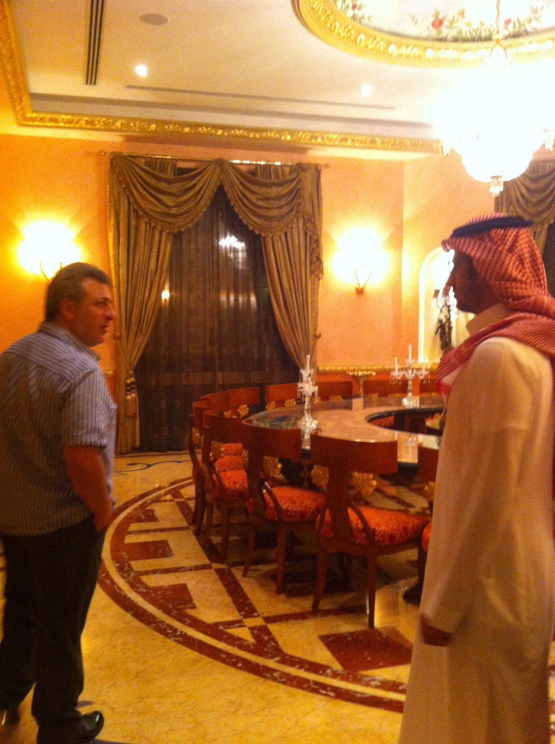 Two men in an ornate dining room. One in Western clothing, the other in traditional Arab attire. A large table and chandelier are visible.