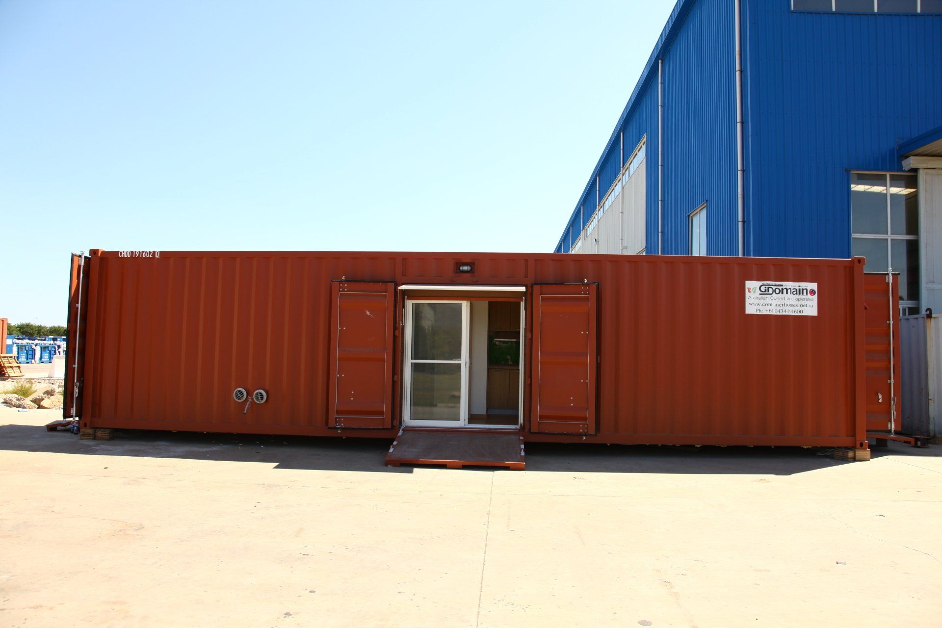 Red shipping container office with open glass door and small ramp. Blue building in background.