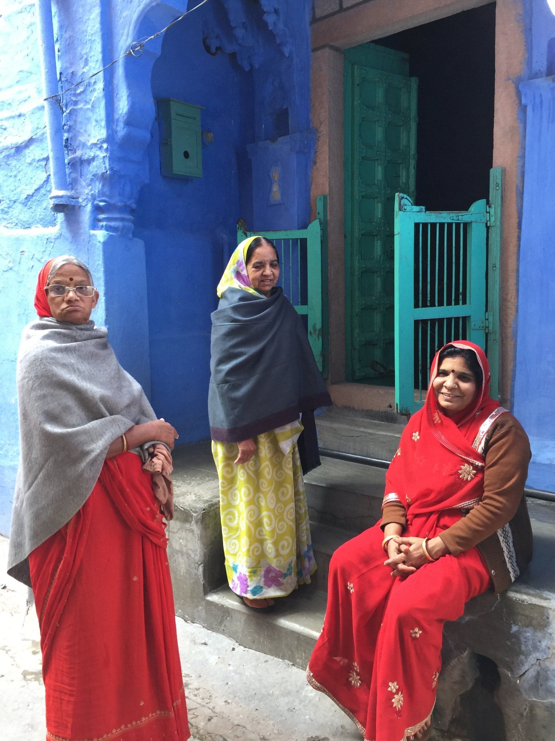 Three women in colorful clothing pose in front of a blue building with green door.