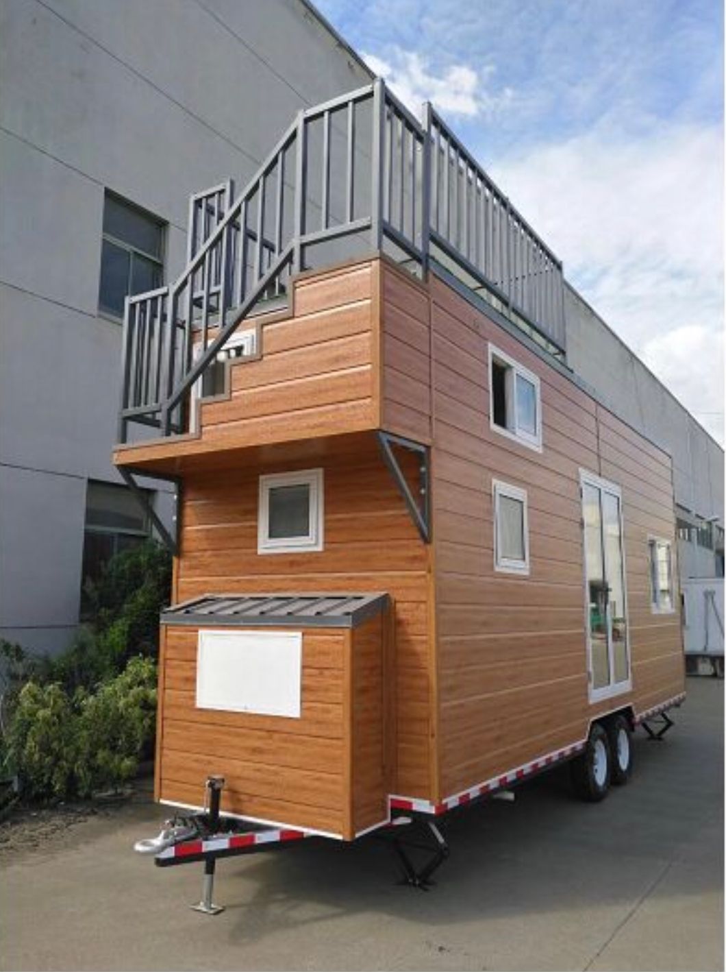 Brown tiny house on wheels with a rooftop deck and stairs, parked outside a building.