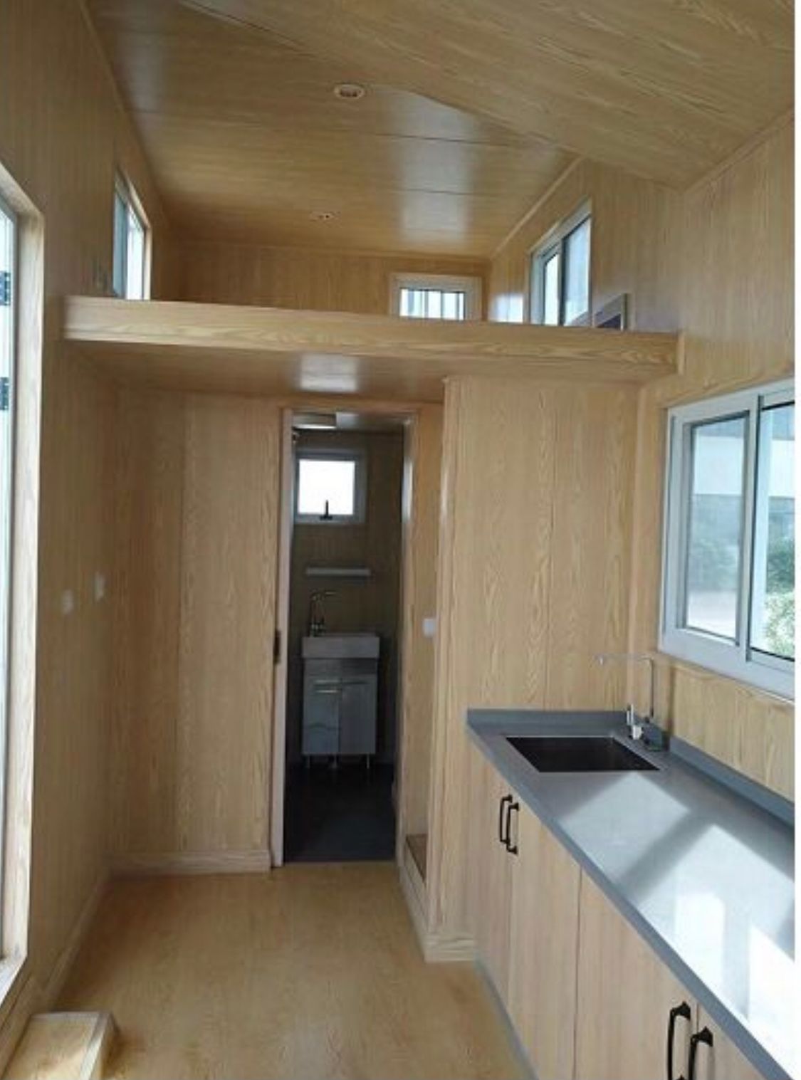 Interior of a tiny house with a kitchen, bathroom, and loft. Light wood paneling, windows, and a black countertop are visible.