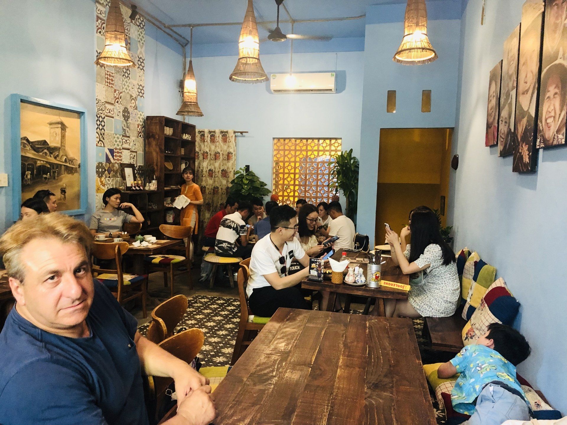 Man in a cafe looks at the camera. People are seated at tables in a blue-walled room, lit by hanging lamps.