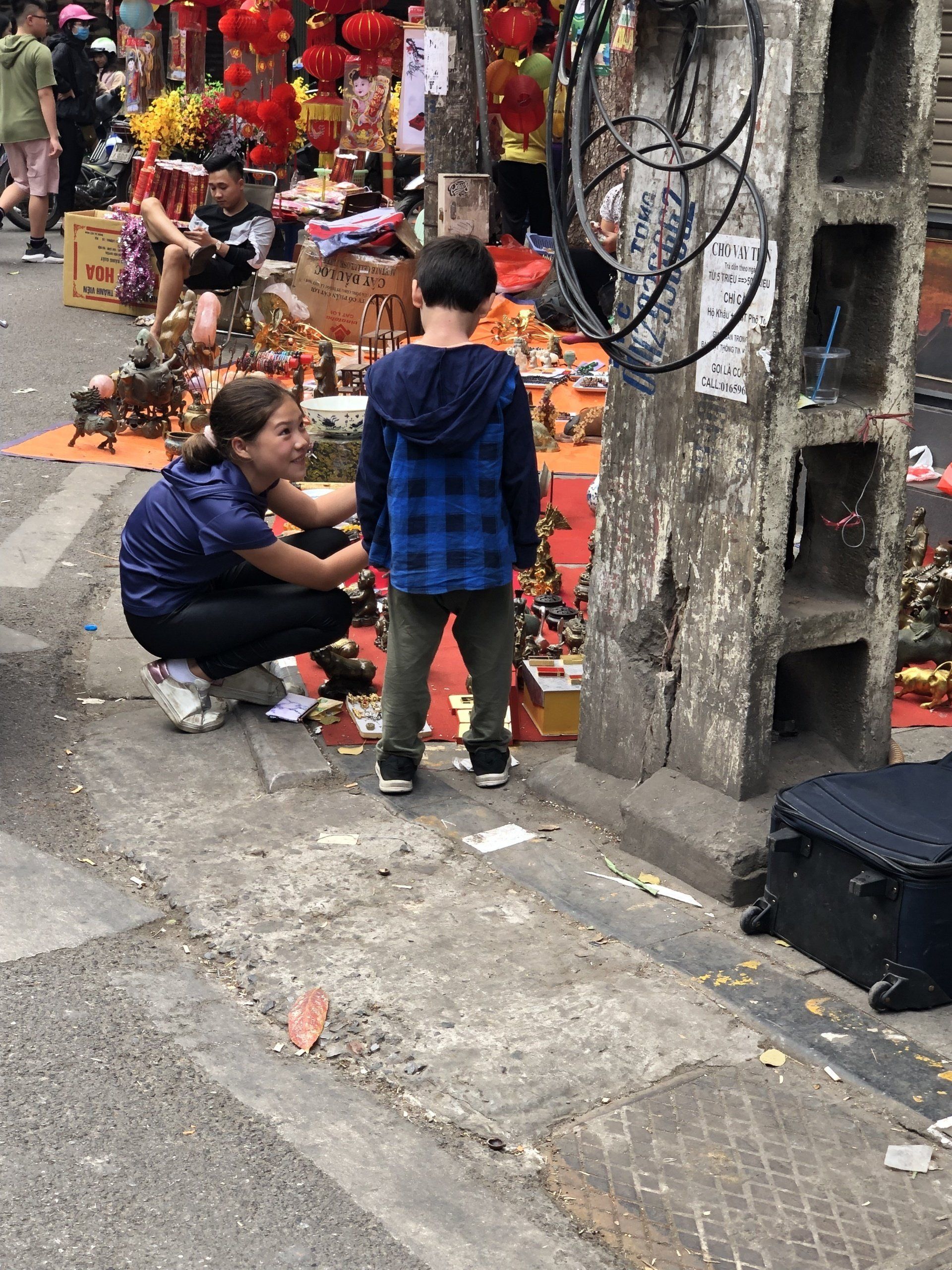 Woman adjusts a child's collar near a display of goods in an outdoor market setting.