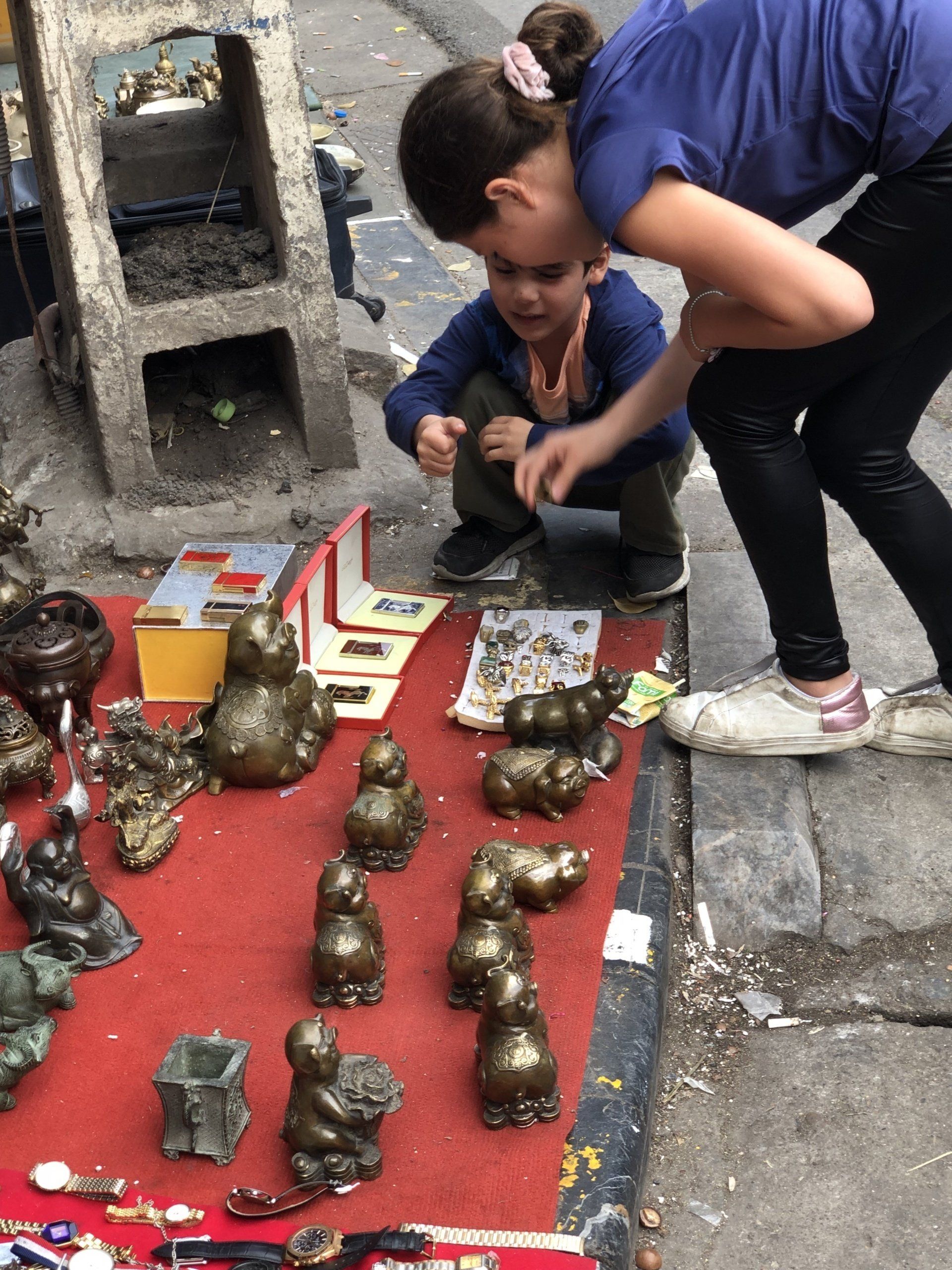 Two people examining small bronze statues at an outdoor market stall.