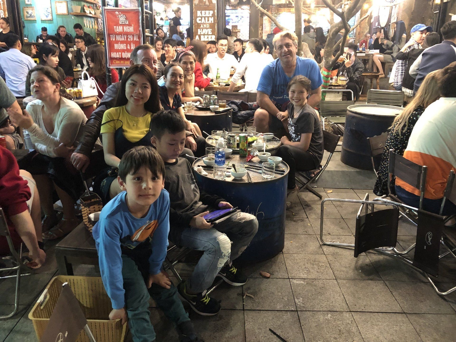 People seated at outdoor tables, drinking in a bustling street scene at night.