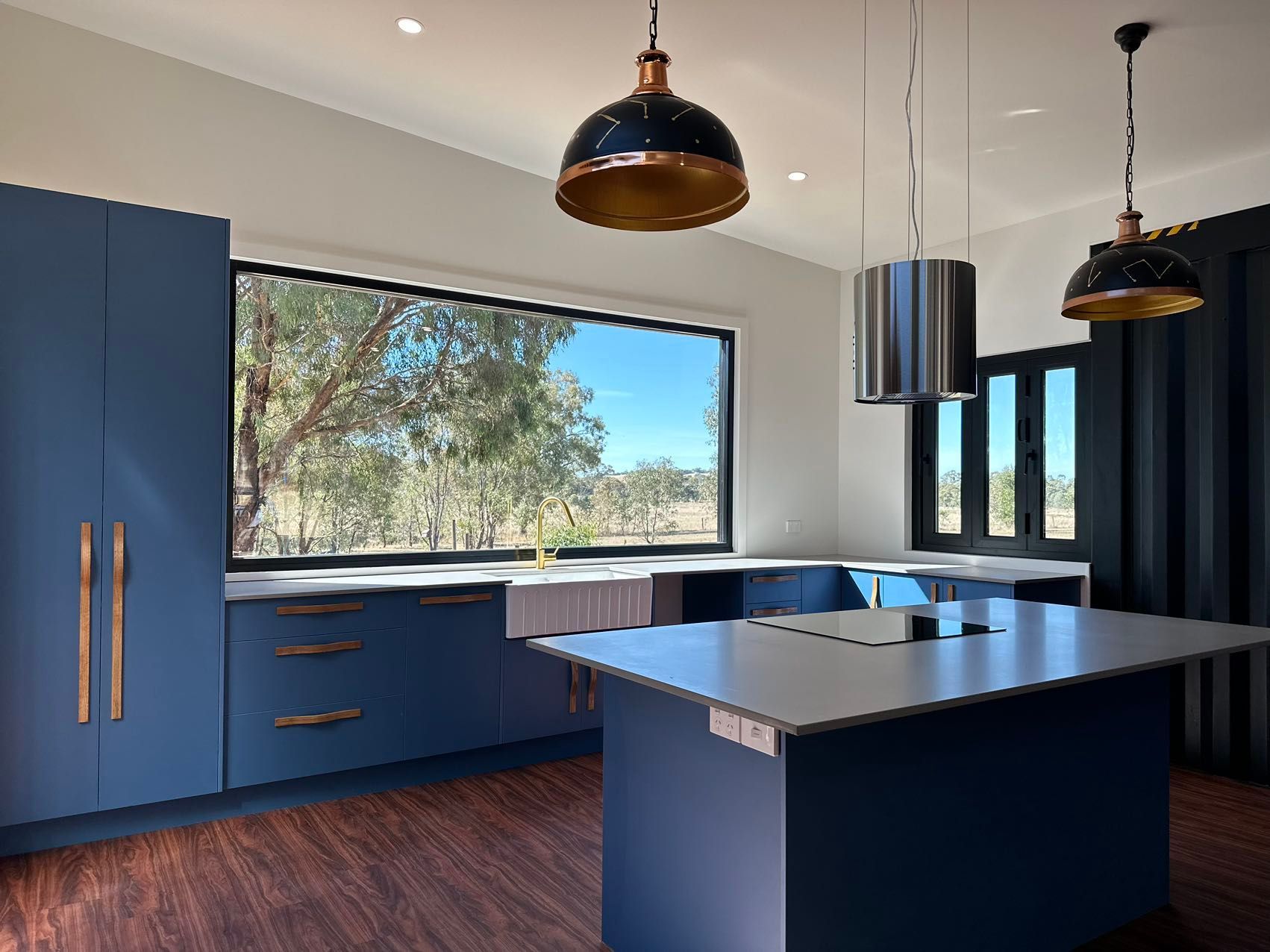 Blue kitchen with island, large window overlooking trees, black and copper pendant lights.