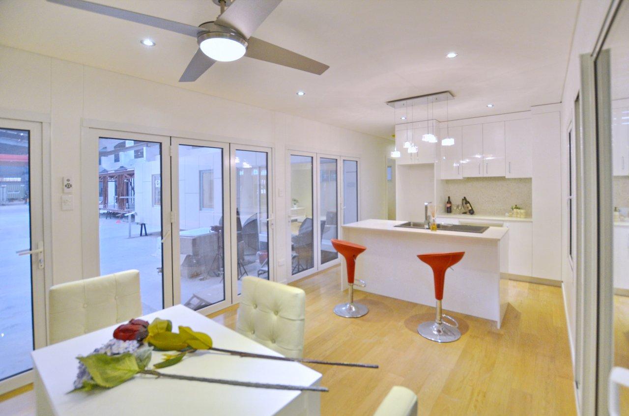 Modern white kitchen with island, red bar stools, and dining table; large windows.
