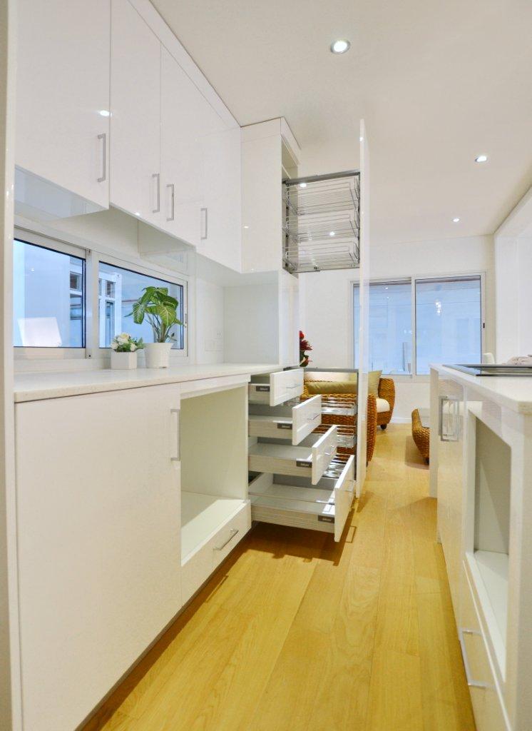 Modern white kitchen with pull-out drawers and wooden floor. Bright, clean design with upper cabinets and a window.